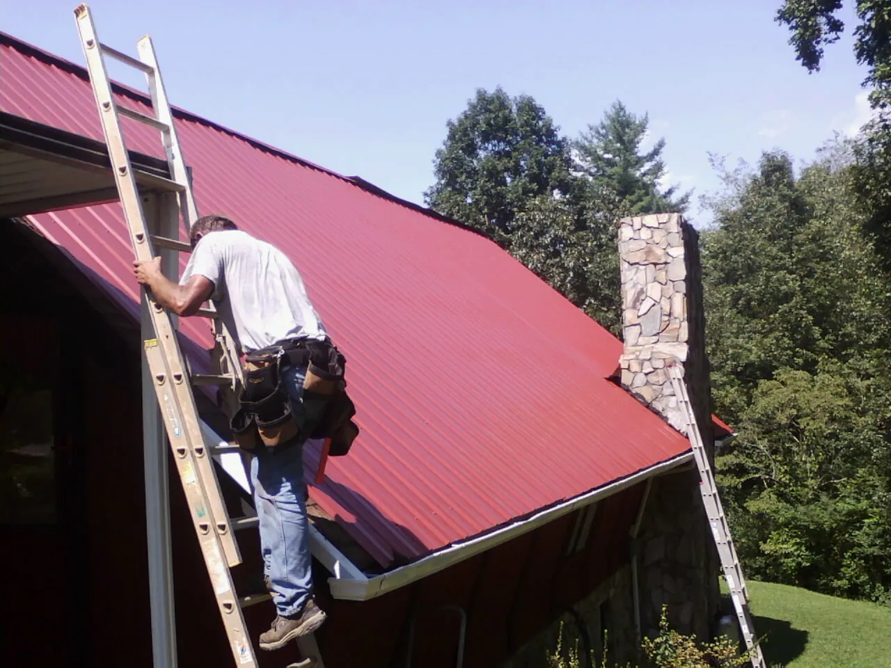 Roofer on a ladder beside a red metal roof and stone chimney — Soffit & Fascia Repair by Roofing Brothers in Spencerport